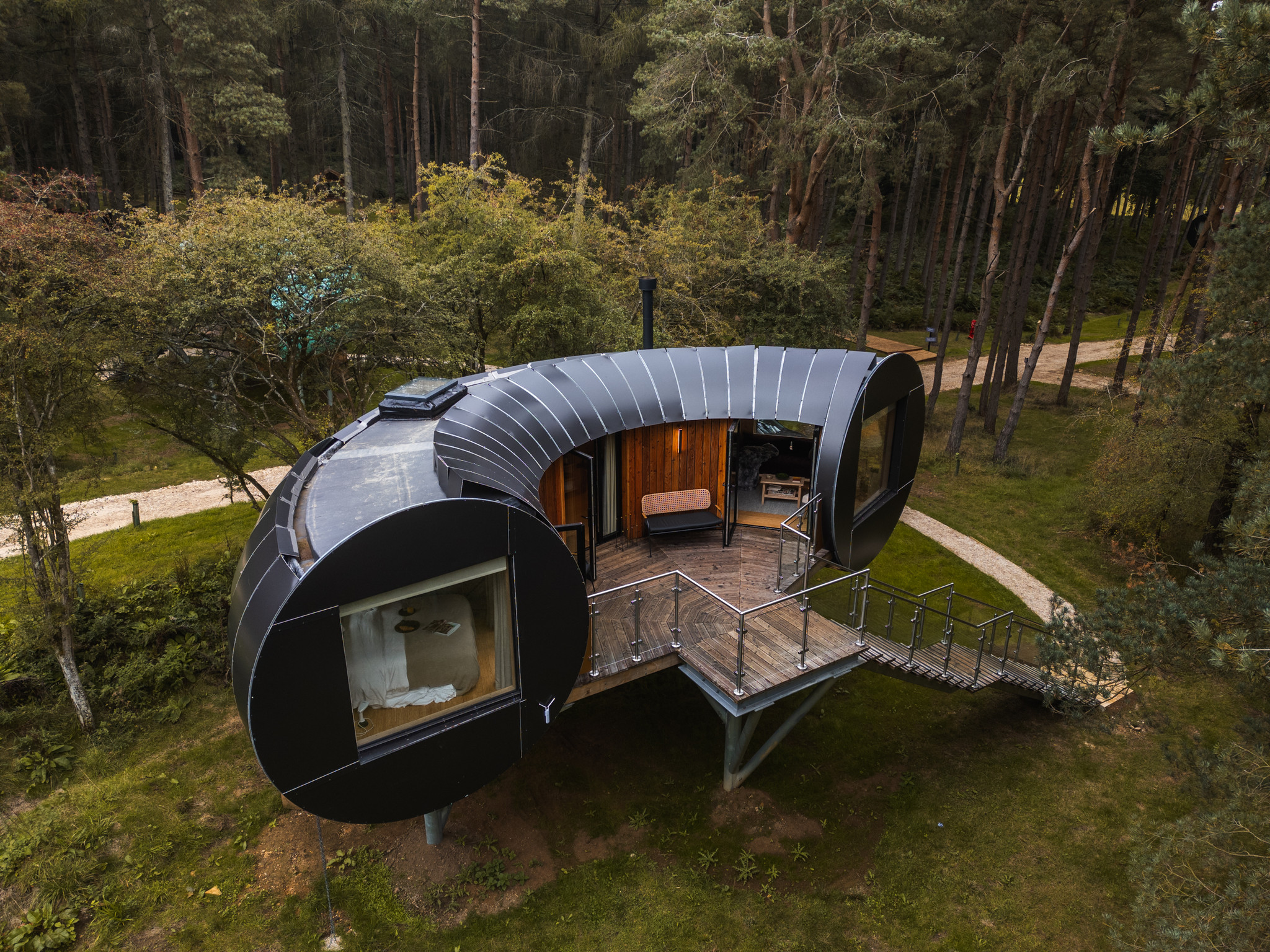 Aerial Majesty: Bastard Balm in the Heart of Nature | TreeDwellers Soar above the enchanting Bastard Balm treehouse with this breath-taking aerial shot, capturing the seamless integration of modern architecture within the lush landscape. Nestled among the towering trees, this exterior view highlights the unique structure and design that make Bastard Balm a standout retreat in the Cotswolds. Experience the serene beauty of its natural surroundings, promising an unforgettable escape amidst the treetops.